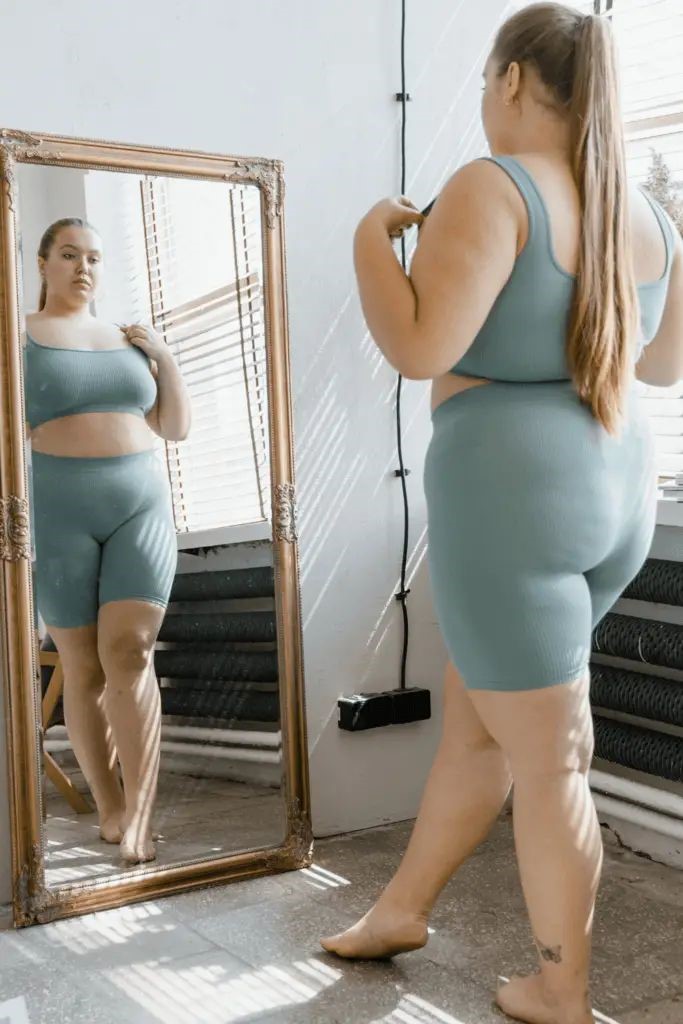 Plus-size woman wearing fitted activewear comparing her body proportions in a full-length mirror.