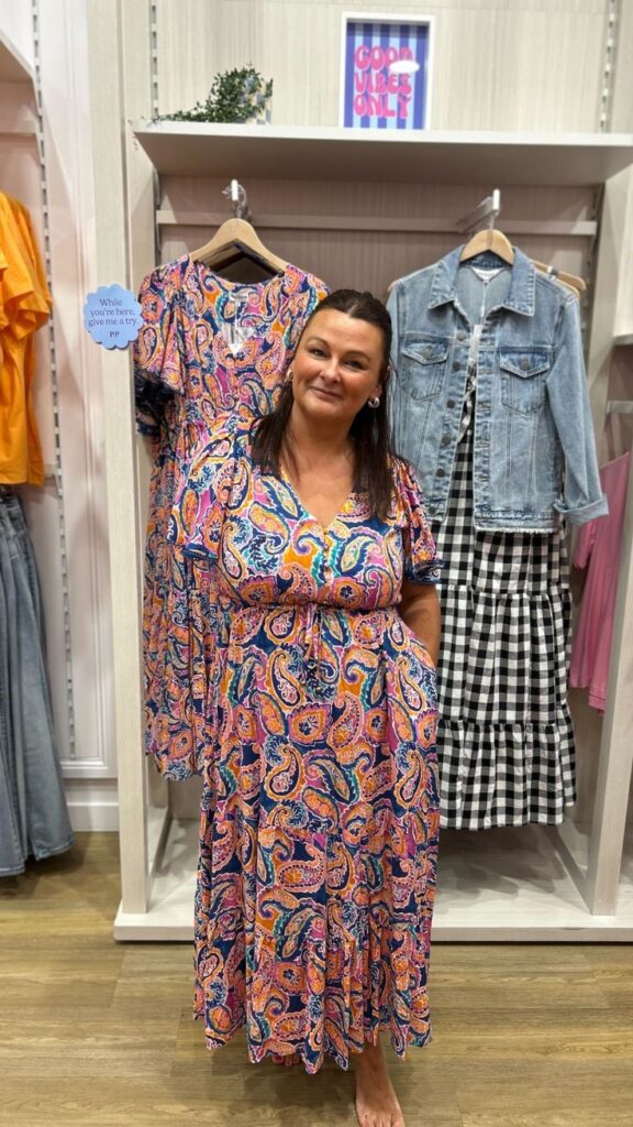 Plus-size woman trying on a colorful paisley maxi dress while shopping in a clothing store.