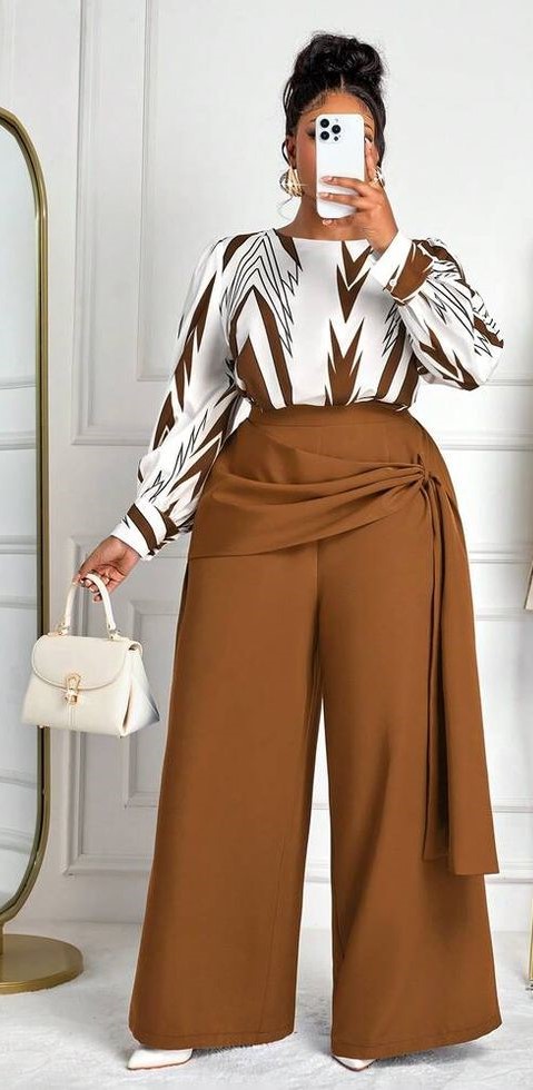 Plus size woman wearing a printed blouse with draped wide-leg trousers, pointed-toe heels, and a white structured handbag.