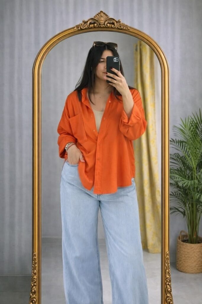 Plus size woman checking the fit of an orange shirt and wide-leg jeans in a mirror 