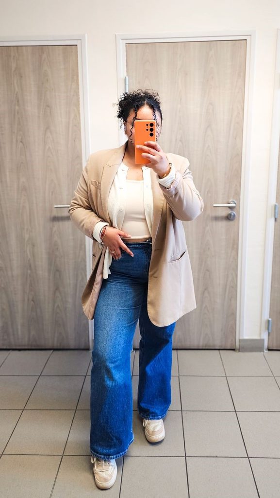 Plus size woman wearing a beige blazer over a white top with blue wide-leg jeans and beige sneakers.