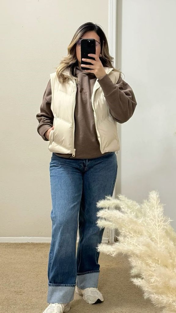 Cozy fall outfit for curvy women featuring a brown hoodie, cream puffer vest, wide-leg blue jeans, and white sneakers