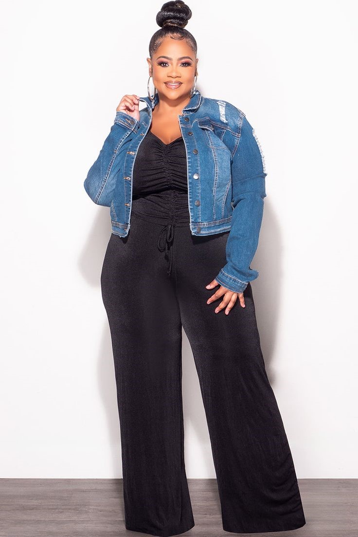 Plus size denim jacket outfit with a black jumpsuit, distressed blue denim jacket, and statement hoop earrings.