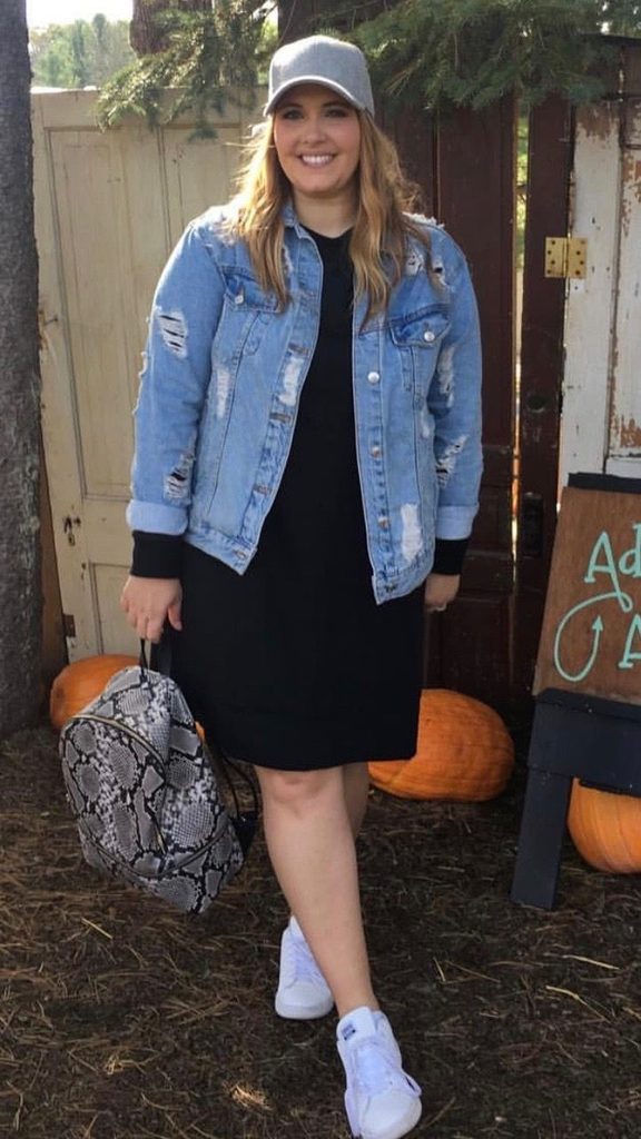 Plus size denim jacket outfit with a black shirt dress, distressed blue denim jacket, and white sneakers.