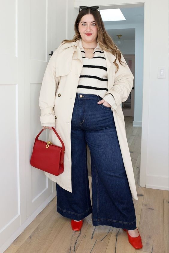 Plus size fall outfit with a cream trench coat, black-and-white striped top, dark-wash wide-leg jeans, and red shoes.