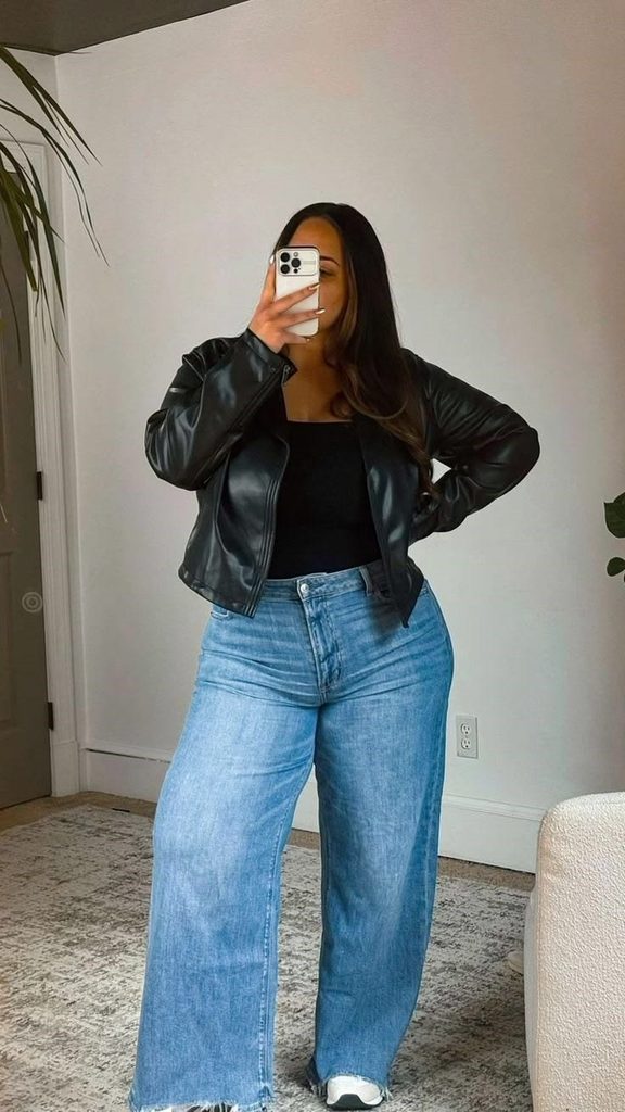 Plus size leather jacket outfit ideas with a black jacket, black top, and wide-leg blue jeans