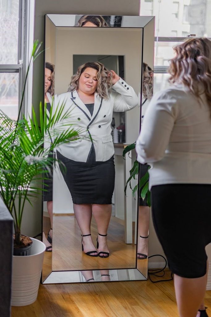 Plus size woman checking her white blazer outfit in the mirror while learning how to tell if you're wearing the wrong size